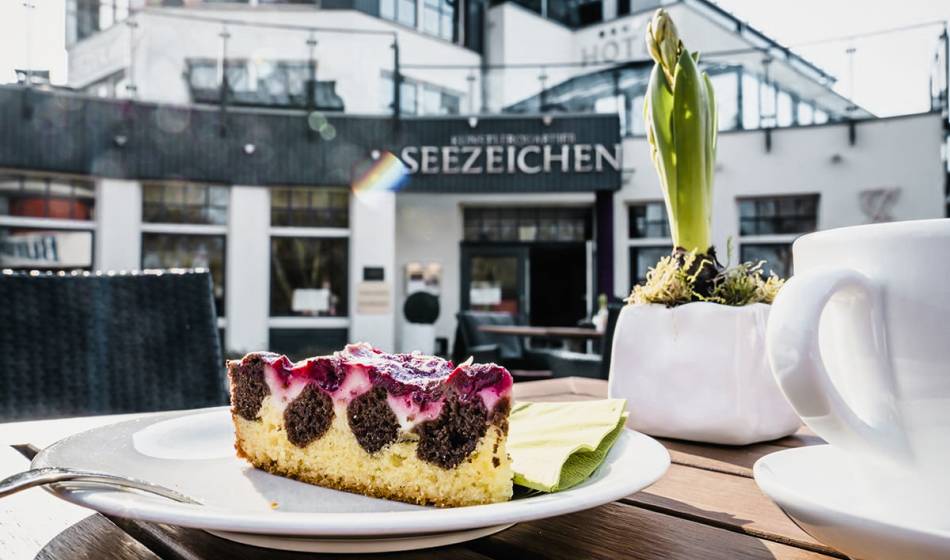 Piece of cake with coffee on a plate, served on the terrace of Café #fraukorn in the Hotel Künstlerquartier Seezeichen.