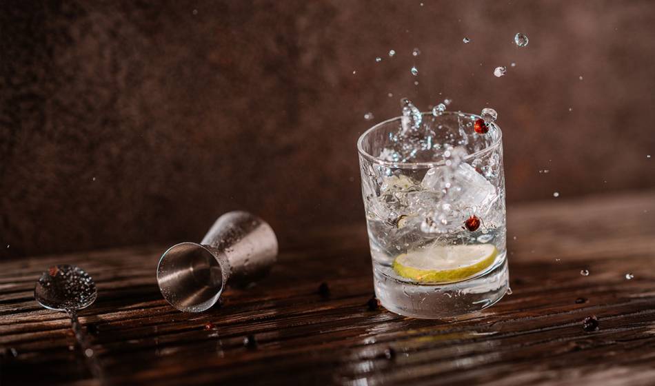 A glass with a clear drink, ice cubes, lemon slice and juniper berries, water splashes out of the glass in #herrhoshi.