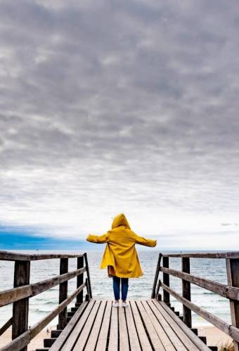 Eine Frau in einer gelben Regenjacke steht mit ausgebreiteten Armen auf einem Holzsteg am Strand von Sylt und blickt auf das Meer