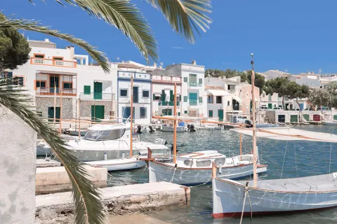 Anlegestelle in Portocolom mit kleinen Booten und farbenfrohen Hausfassaden mit bunten Fenstern und Türen im Hintergrund.