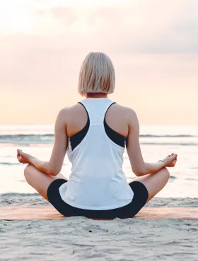Eine Frau sitzt meditierend am Strand, der Blick auf das Meer gerichtet, während die Sonne aufgeht