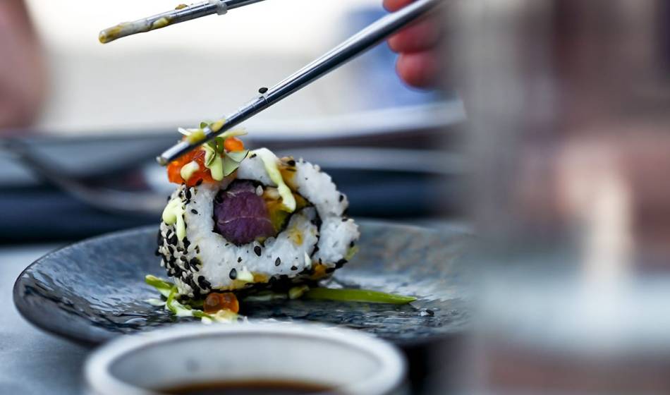 Close-up of a California roll with salmon, avocado and sesame held by chopsticks over a plate at #herrhoshi.