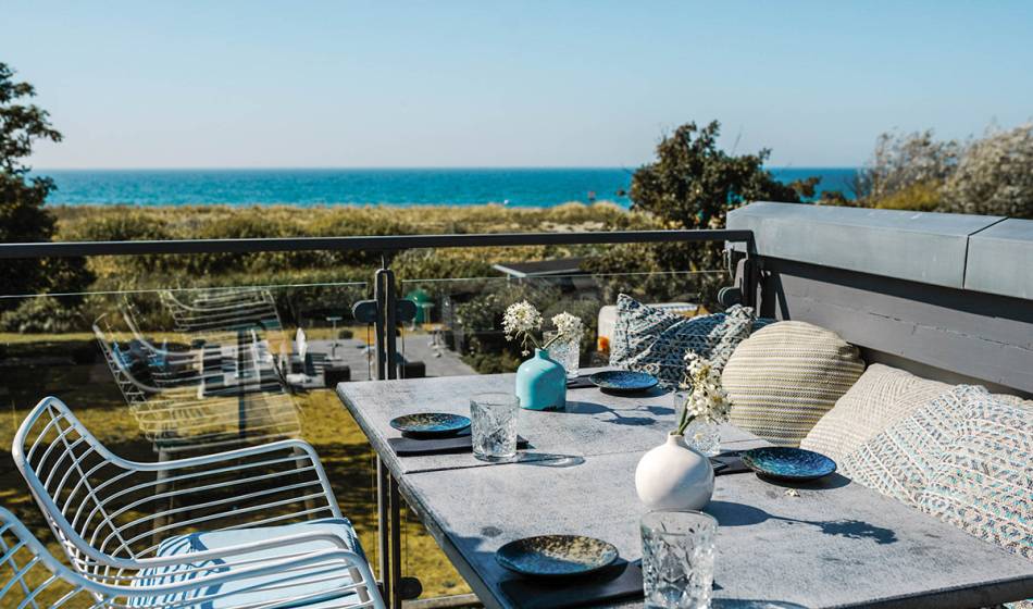 Stylish terrace at #herrhoshi in Ahrenshoop with tables, cushions and glass railings, offering a view of the Baltic Sea.