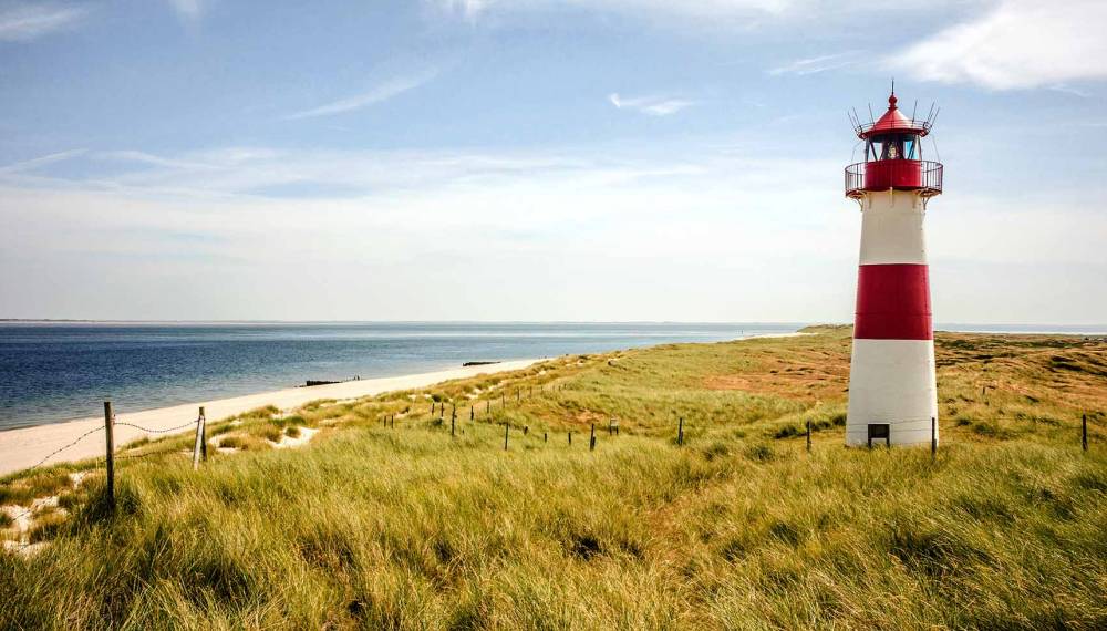 Ein rot-weißer Leuchtturm steht inmitten der Dünenlandschaft auf Sylt mit Blick auf die Nordsee.
