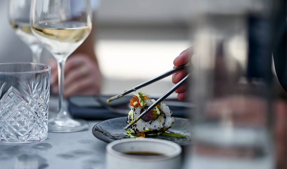 Close-up of inside-out sushi with sesame and vegetable filling, held by chopsticks, next to wine glasses in #herrhoshi.