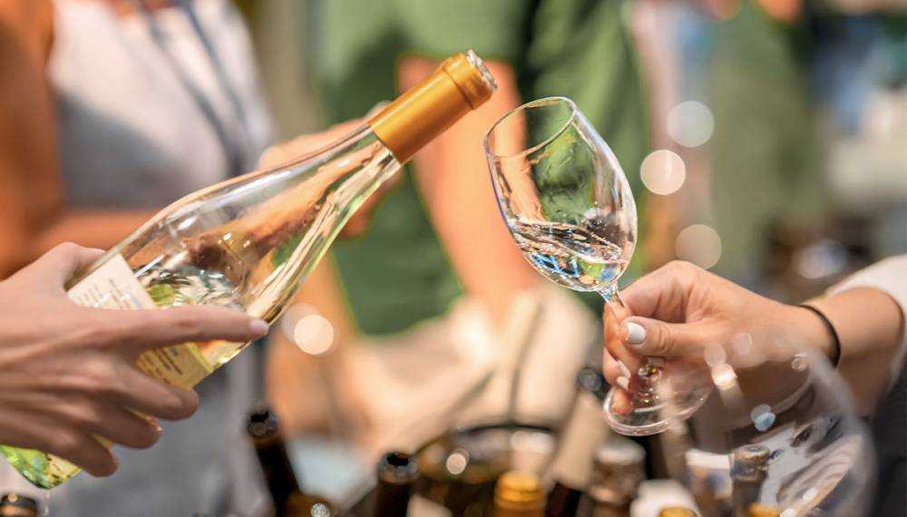 Weißwein wird aus einer Flasche in ein Weinglas eingeschenkt, im Hintergrund stehen weitere Weinflaschen und Personen.