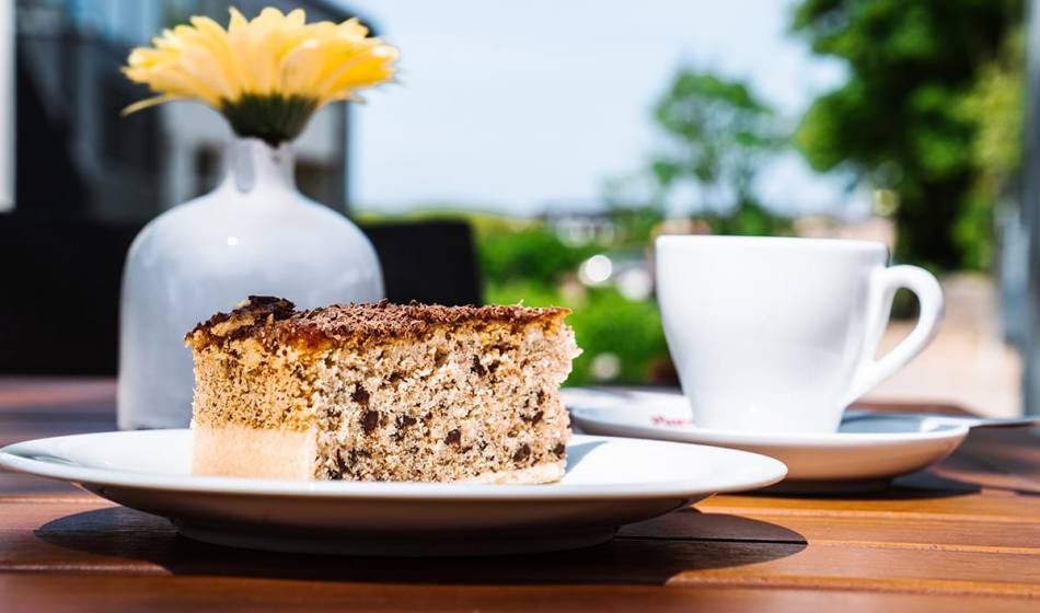 Closeup of cake on white plate with tea cup in #fraukorn outdoors on wooden table with a flower in the background.