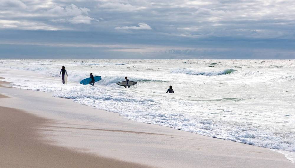 Surfer mit Brettern stehen am Weststrand von Sylt in der Brandung der Nordsee unter bewölktem Himmel.