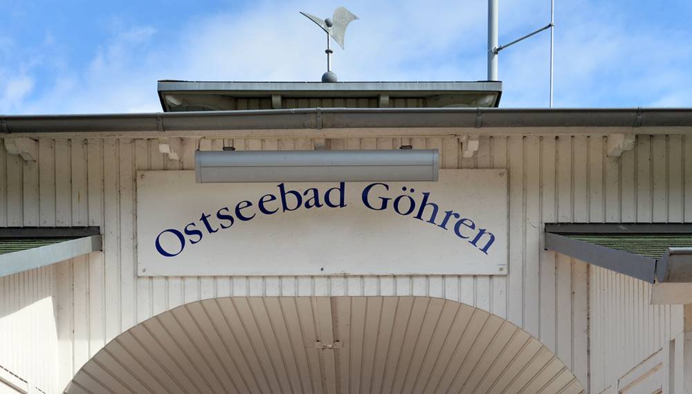 Ortsschild „Ostseebad Göhren“ am Eingang der Seebrücke mit weißem Holz und blauem Himmel.