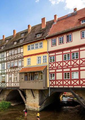 Porträt der Erfurter Krämerbrücke mit bunten Fachwerkhäusern, Kirchturm und Grünfläche am Wasserlauf in Thüringen