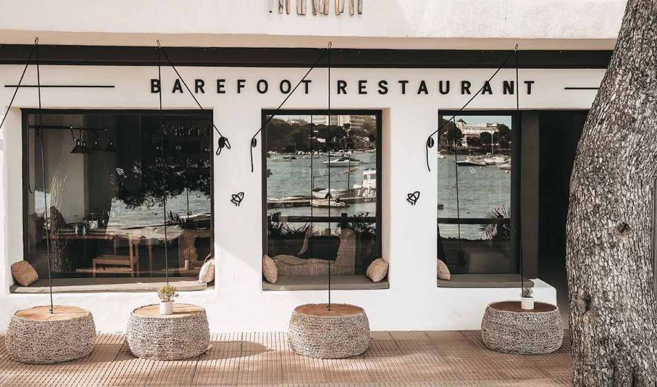 Exterior view of the Barefoot Restaurant Mallorca with comfortable seating and panoramic views of the harbor through the large windows.