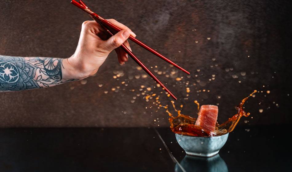 A tattooed hand with chopsticks holds sashimi over a bowl of sauce that splatters when dipped in #herrhoshi.