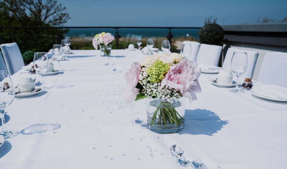 Outdoor wedding table with white tablecloth, glass vases with flowers and Baltic Sea view in the background.