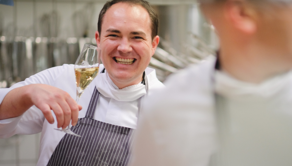 Johannes Wallner lacht und prostet mit Champagnerglas in der Hotelküche des Hotel Elephant in Weimar beim Event Wallners Lieblinge.