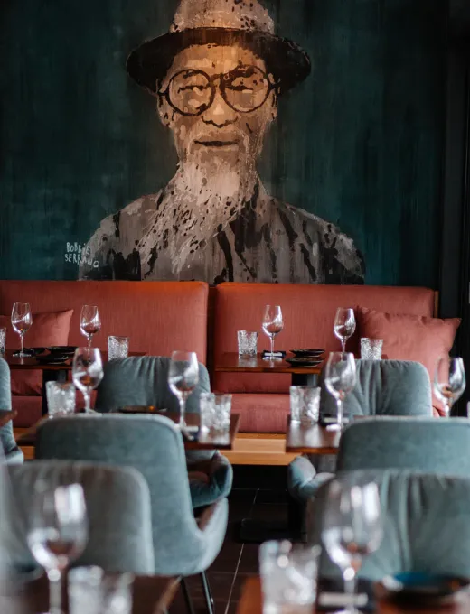 Restaurant mit gedeckten Tischen, Weingläsern und einer großen Wandmalerei eines Mannes mit Hut und Brille im Hintergrund.