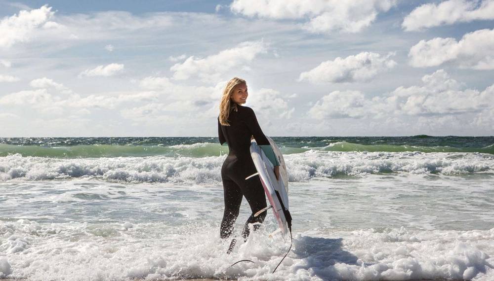 Eine Frau steht mit einem Surfboard im Meer an der Nordsee und blickt Richtung Wellen.