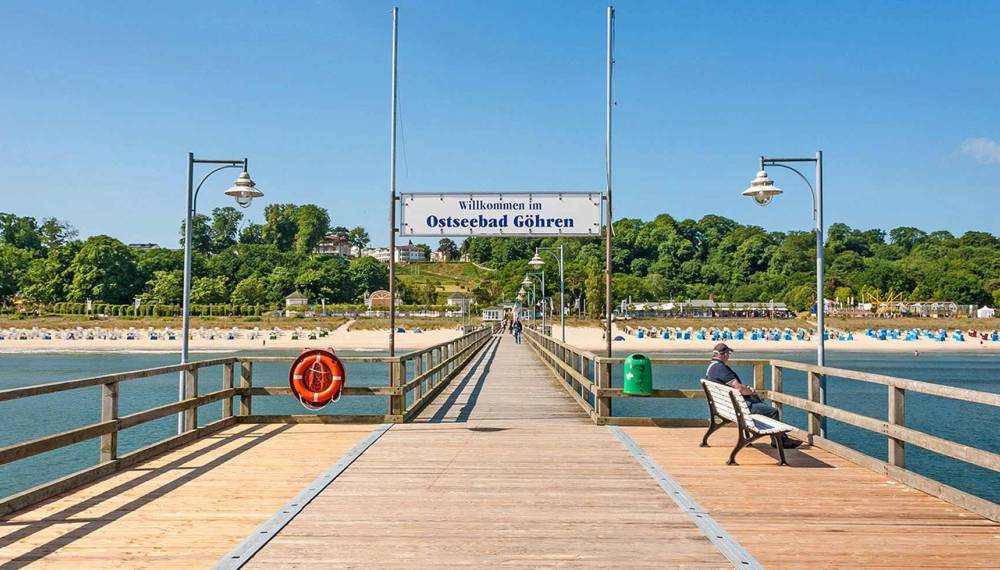 Seebrücke Göhren mit Begrüßungsschild und Blick auf den Strand mit Strandkörben und grüner Küstenlandschaft.