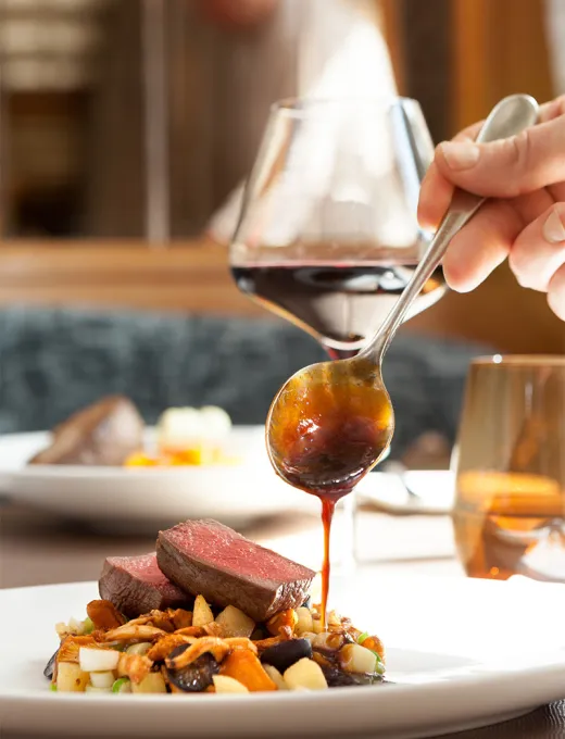Hand eines Kochs, der eine Sauce über ein Stück Fleisch auf einem Teller mit Gemüse gibt, dazu ein Glas Rotwein auf dem Tisch.
