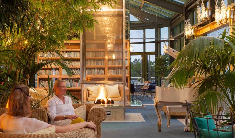 People sit in the winter garden of the Kaiserhof Usedom, surrounded by rattan furniture, a fireplace and bookshelves.