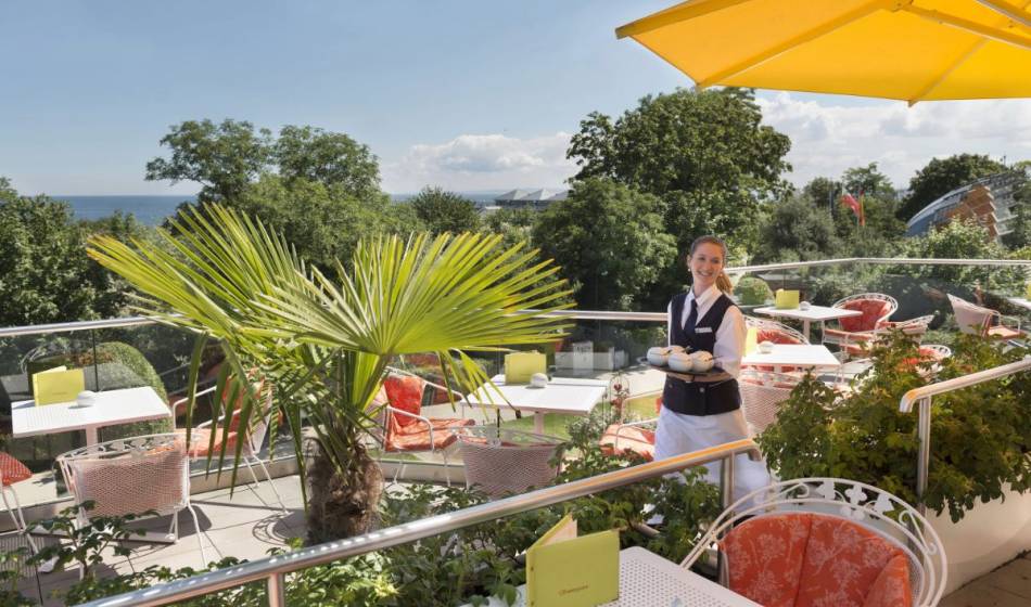 Sunny terrace of the summer garden at Kaiserhof Heringsdorf on Usedom featuring white tables, red chairs, yellow sun umbrellas, and tropical plants. A waitress carries drinks while trees and the Baltic Sea can be seen in the background.