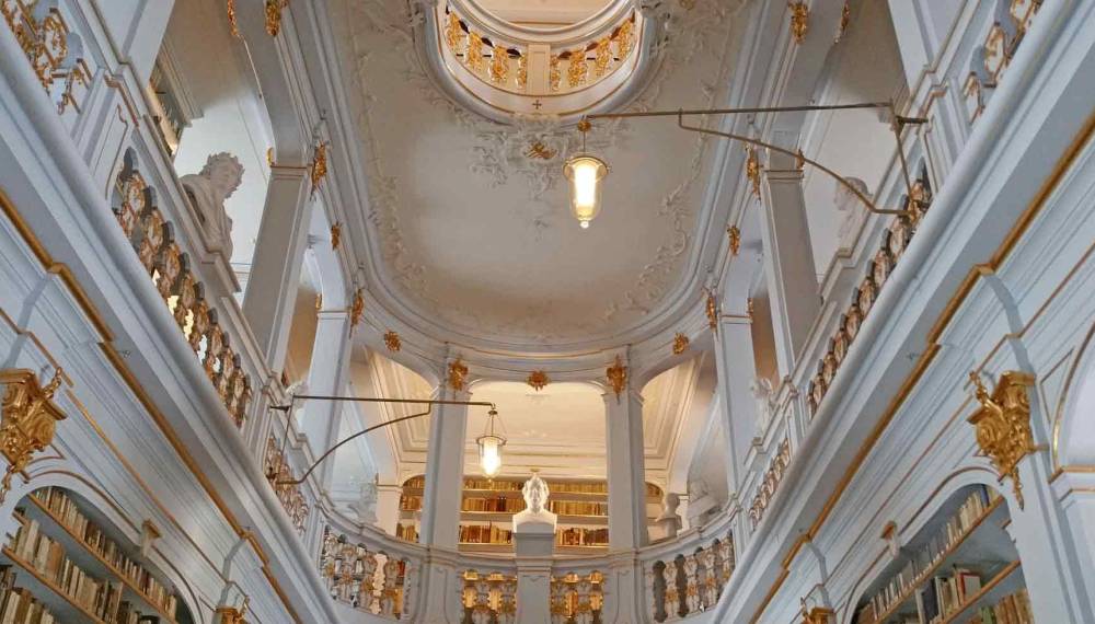Blick in den Rokokosaal der Herzogin Anna Amalia Bibliothek mit umlaufenden Bücherregalen, weißen Säulen, vergoldeten Verzierungen und einer Kuppeldecke.