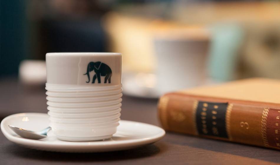 White coffee cup with an elephant logo on a saucer in the Hotel Elephant Weimar, next to a book.