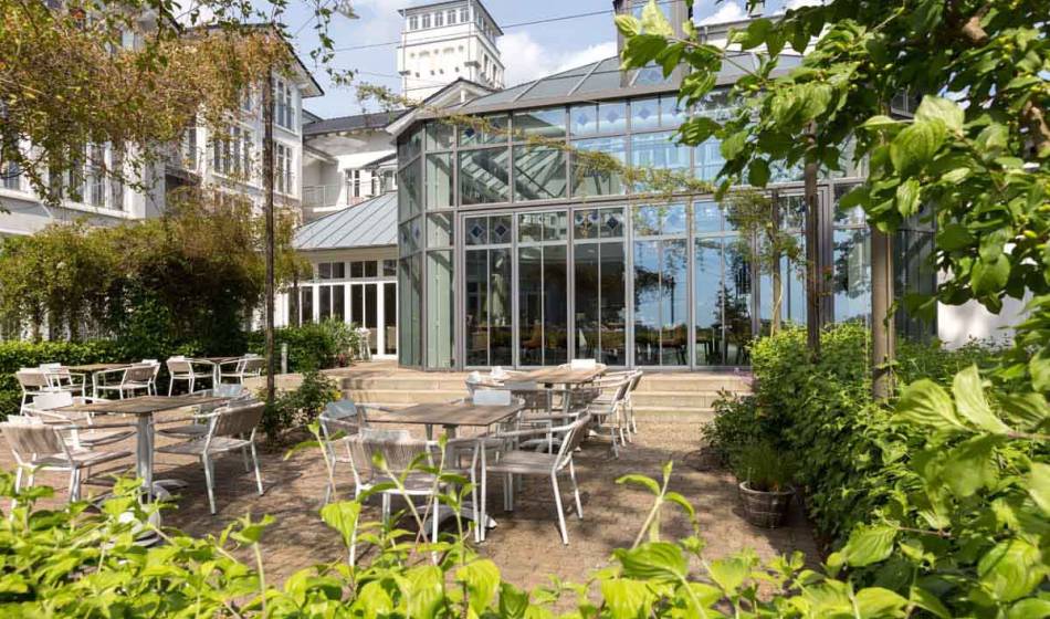 Exterior view of the Strandläufer Restaurant on Rügen with a modern glass facade, surrounded by plants and a furnished terrace.