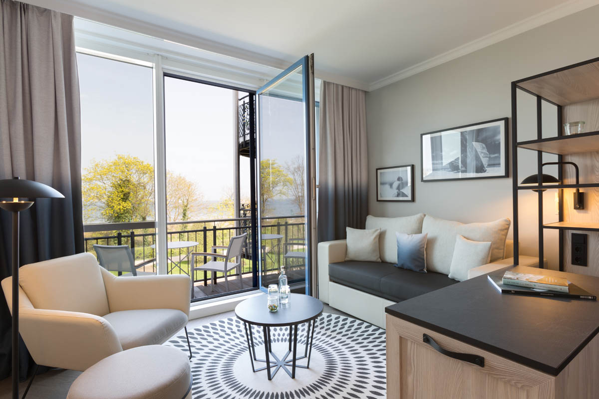 Helles Zimmer mit Sitzbereich, Sofa, Couchtisch und offenem Balkon, umgeben von Bäumen im Hotel Kaiserhof Heringsdorf.