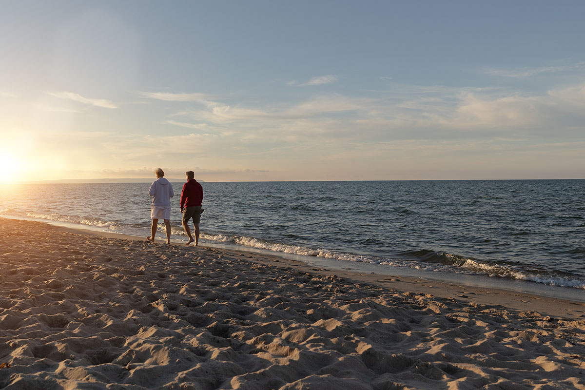 Two people walk barefoot on the beach towards the sunset during their fall vacation at Koopmanns Hotel.