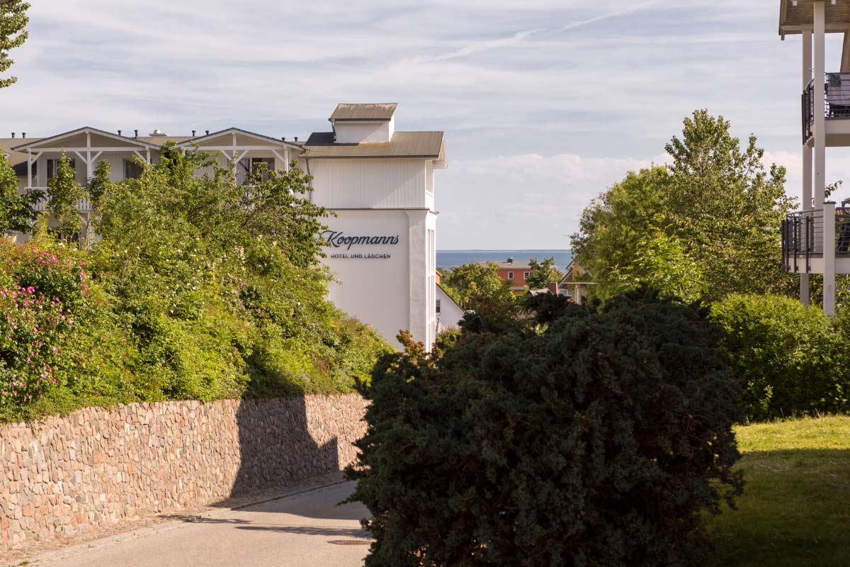 Zugang zum Koopmanns Hotel &amp; Lädchen auf Rügen, umgeben von grünen Büschen und einem Steinweg.