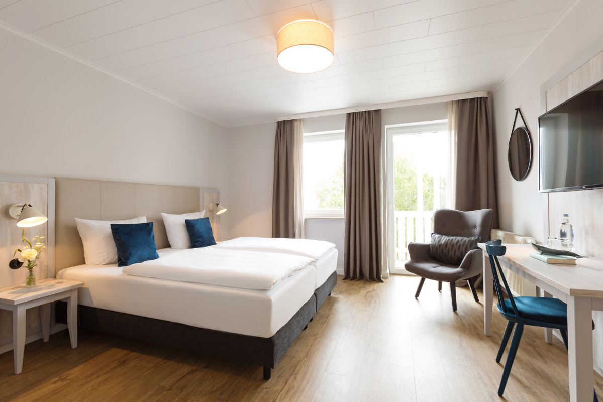 Helles Doppelzimmer im Koopmanns Hotel mit gemütlichem Bett, Balkon und Arbeitsbereich, ideal für erholsame Tage auf Rügen.