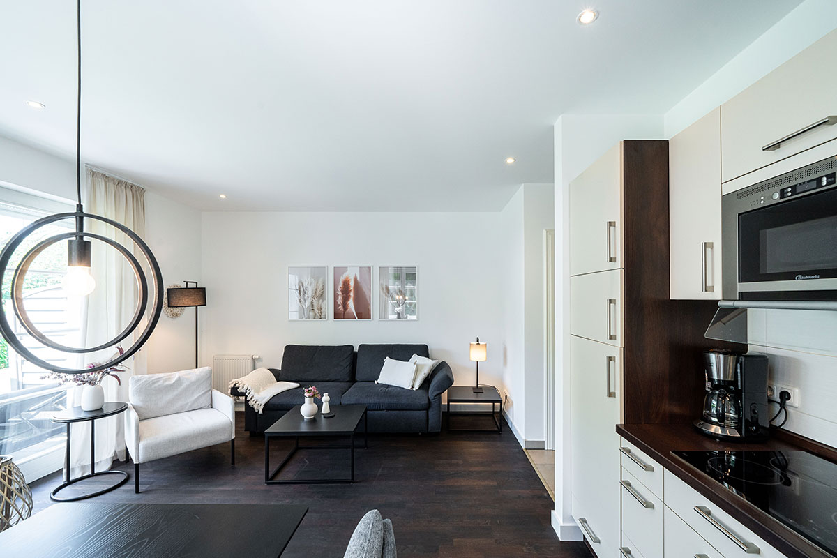 Heller Wohnbereich mit Sofa, Küchenzeile und großen Fenstern in der Ferienwohnung Seeschwalbe im Hotel Künstlerquartier Seezeichen.