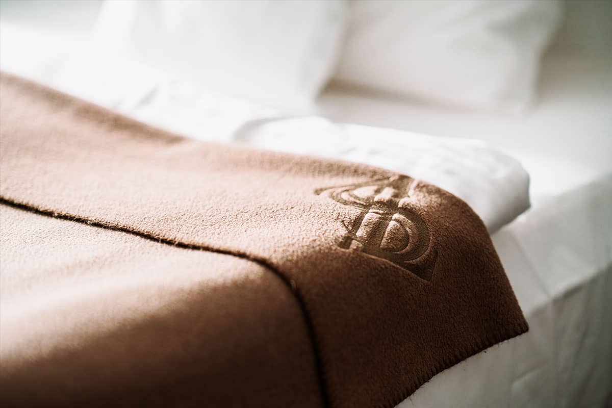 Braune Decke mit Hotel-Logo auf einem Bett im First-Class-Doppelzimmer des Hotel Künstlerquartier Seezeichen.