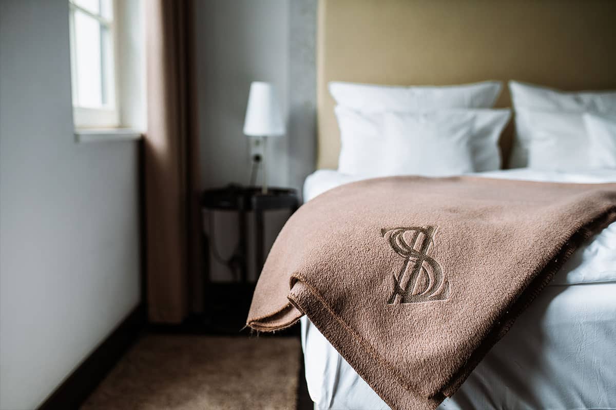 Detailaufnahme einer braunen Decke mit Logo auf einem Bett im First-Class-Doppelzimmer im Hotel Künstlerquartier Seezeichen.