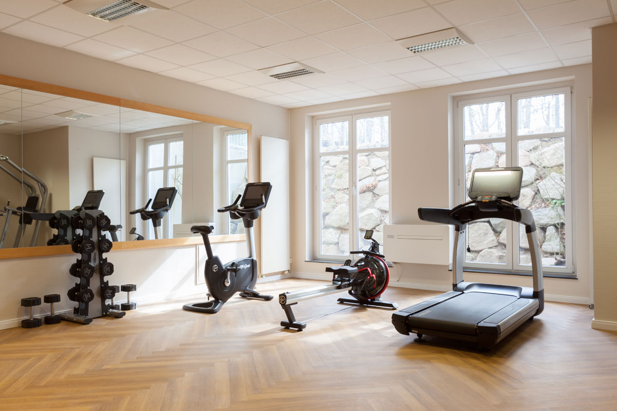 Modern fitness room in the Vju Hotel on Rügen with treadmill, bicycle ergometer, rowing machine and dumbbells in front of large windows.