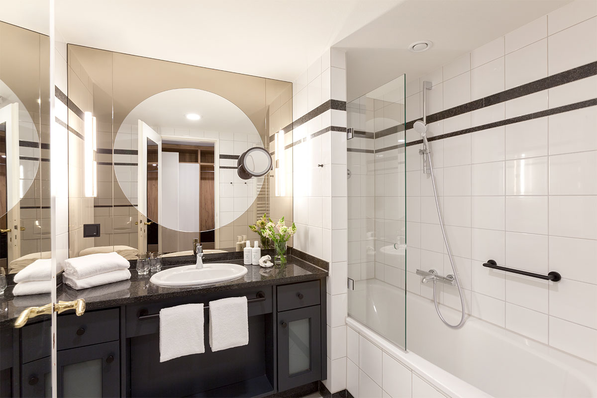 Modernes Hotelbad mit großer Spiegelfläche, Waschbecken, Handtüchern und Badewanne im Einzelzimmer des Vju Hotels.