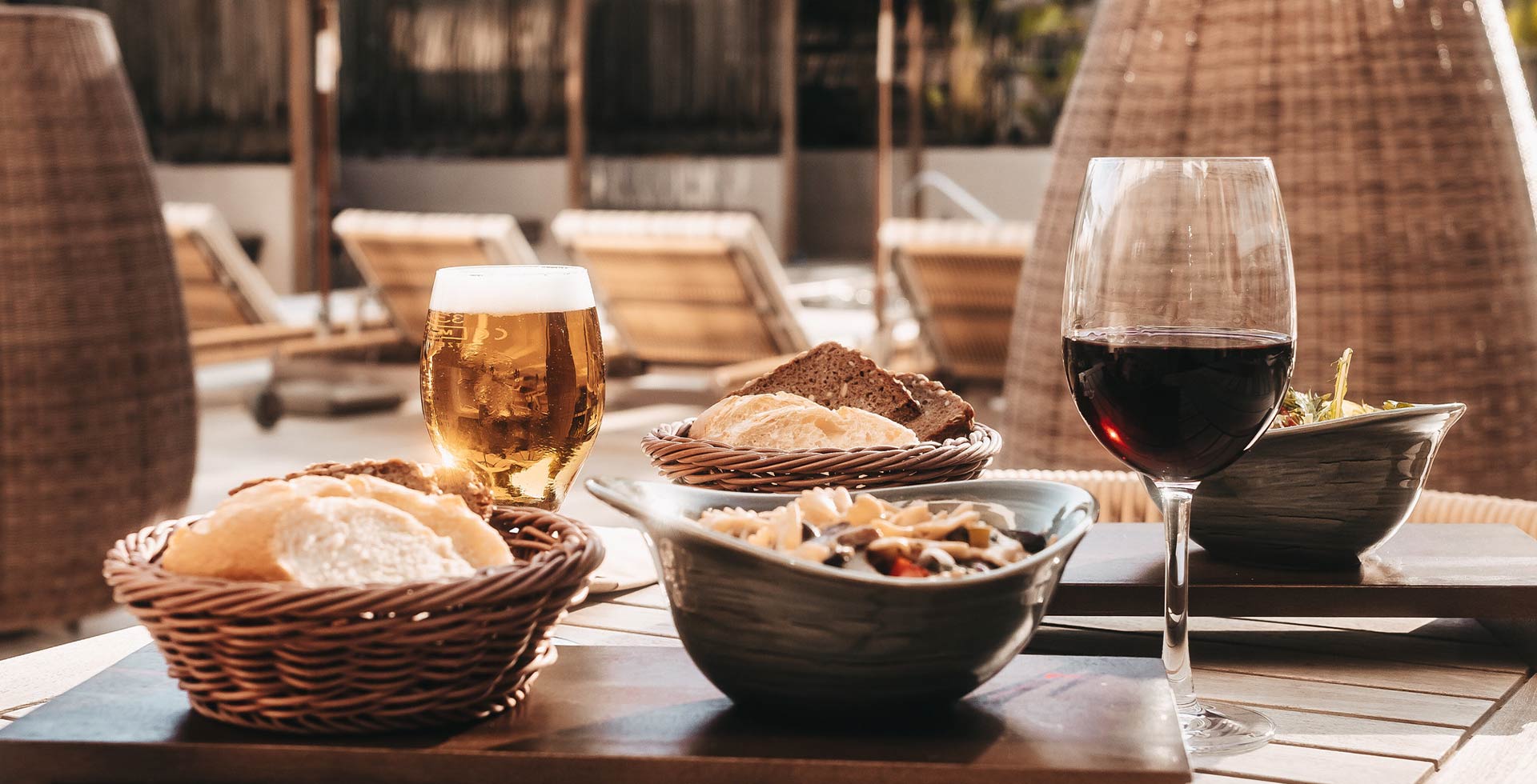 A richly set table with pasta, salad, and bread, accompanied by beer and wine. In the background, wellness loungers in the hotel's outdoor area.