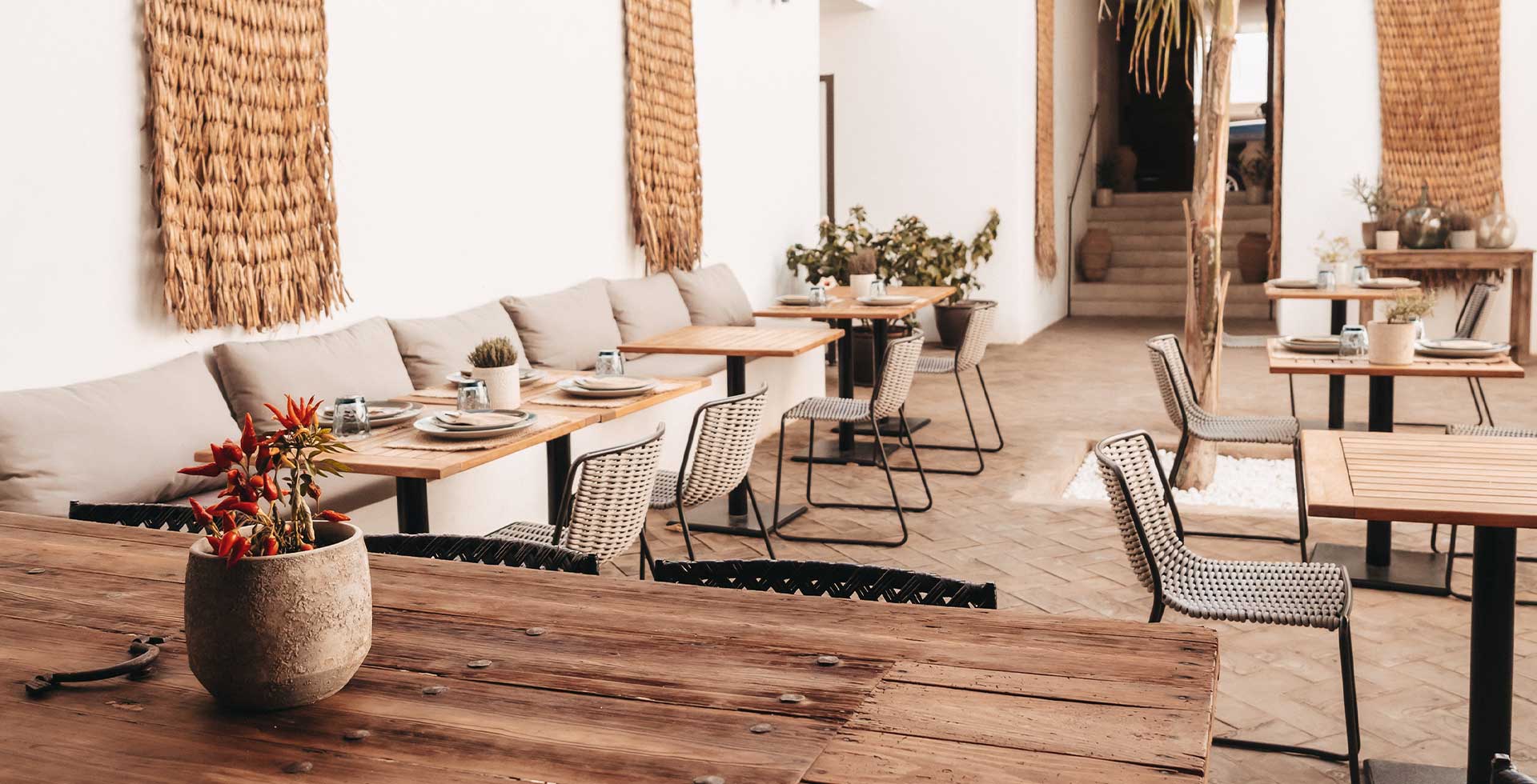 A courtyard decorated with woven art, featuring cushioned benches and minimalist wicker chairs at set wooden tables.