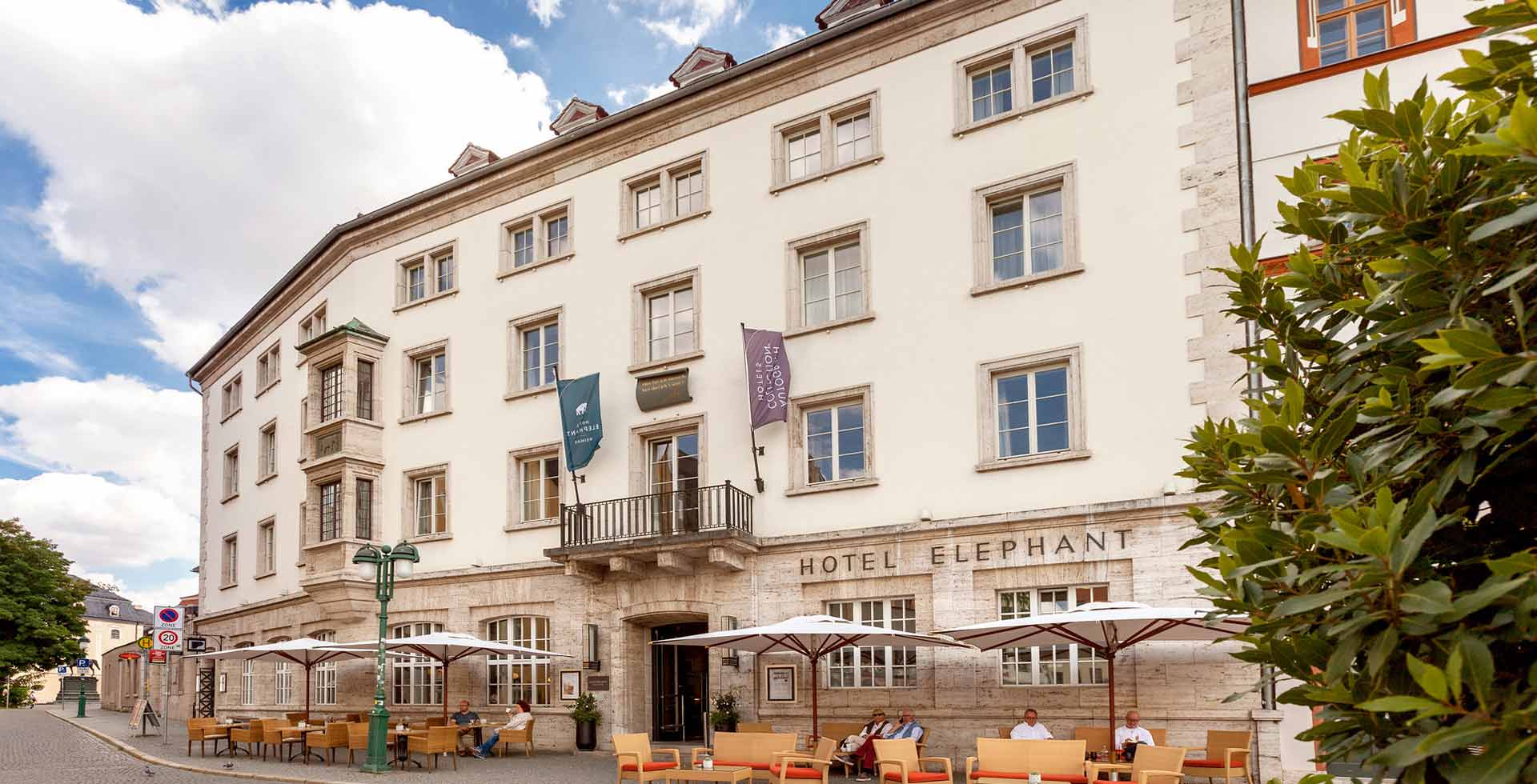 Exterior view of the historic Elephant Weimar hotel building in summer, with comfortable seating areas and large sunshades in front.