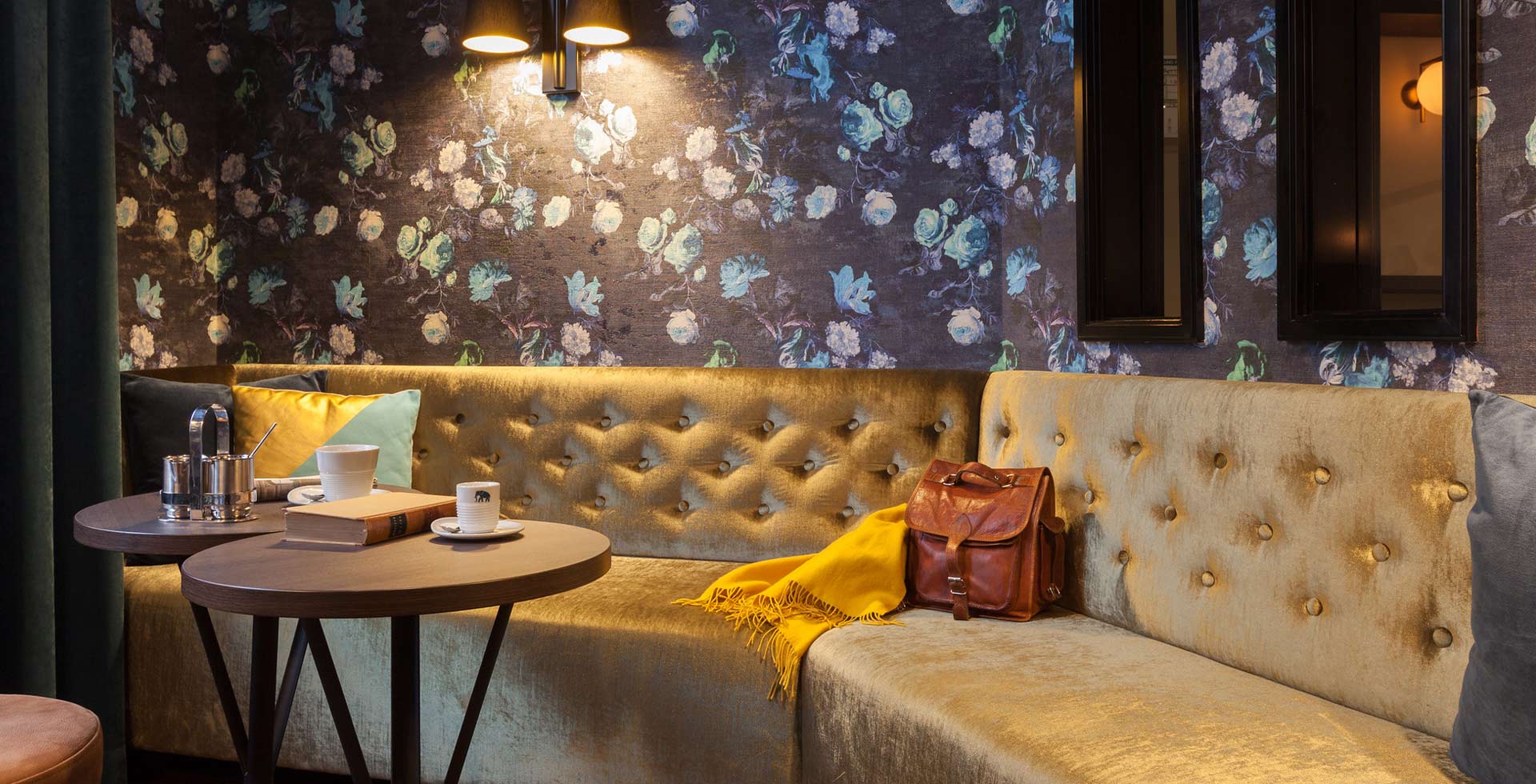 A classic-looking, gold-yellow velvet bench with button tufting in front of blue rose wallpaper in the Weinkost Bar of the hotel.