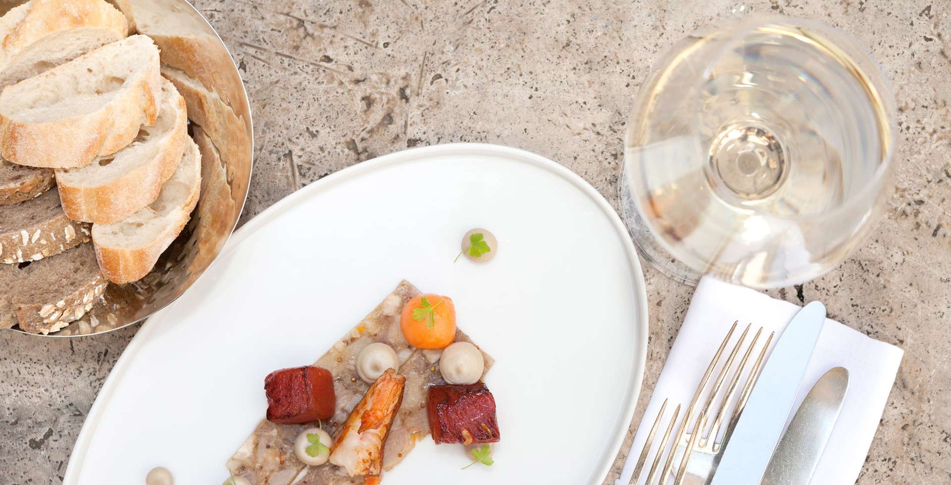 A finely curated meal of fish, seafood, and vegetables with a side of bread and white wine, artfully arranged on a stone table.