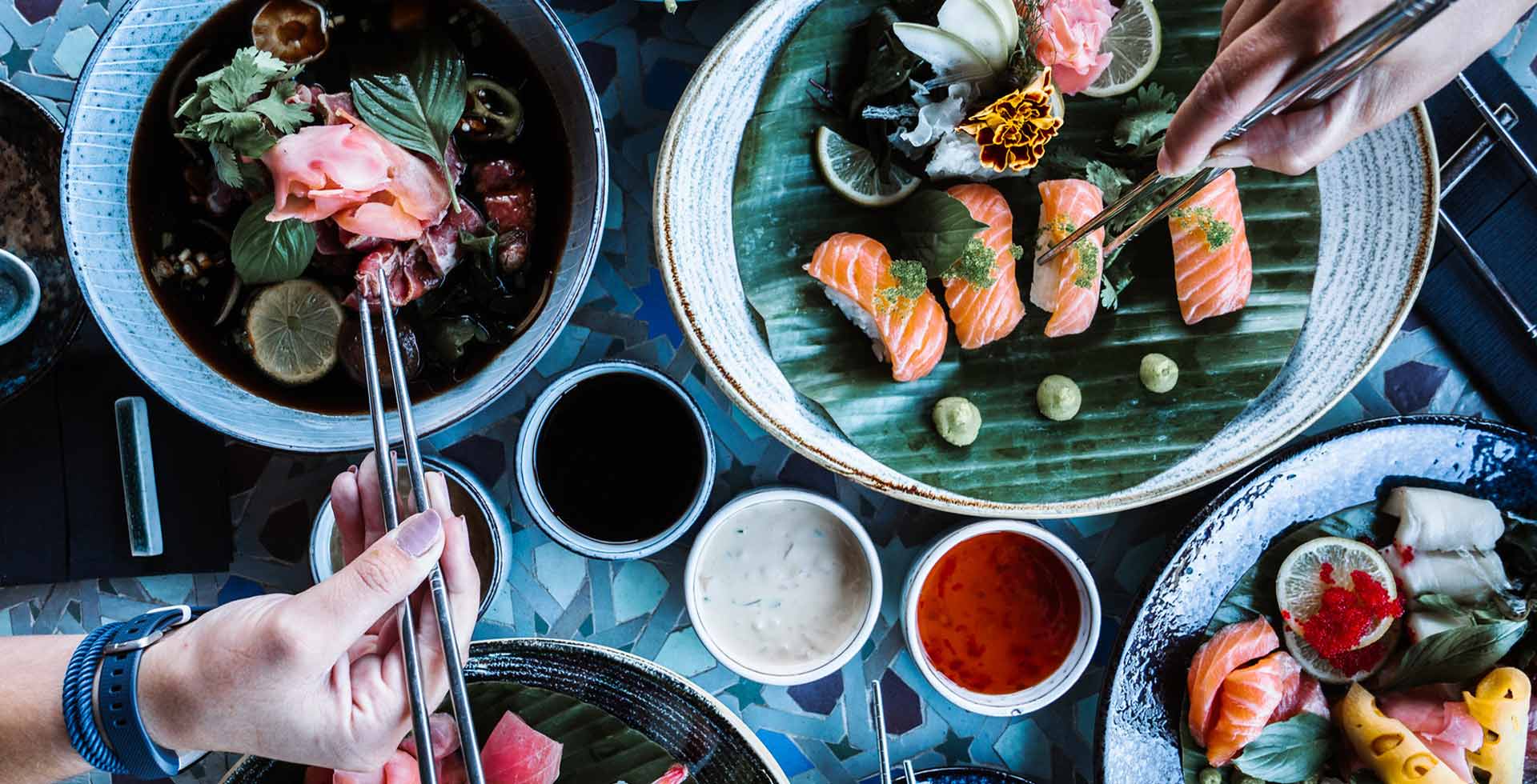 A lavishly set table with sushi and other Asian specialties. Two people are picking food with chopsticks.